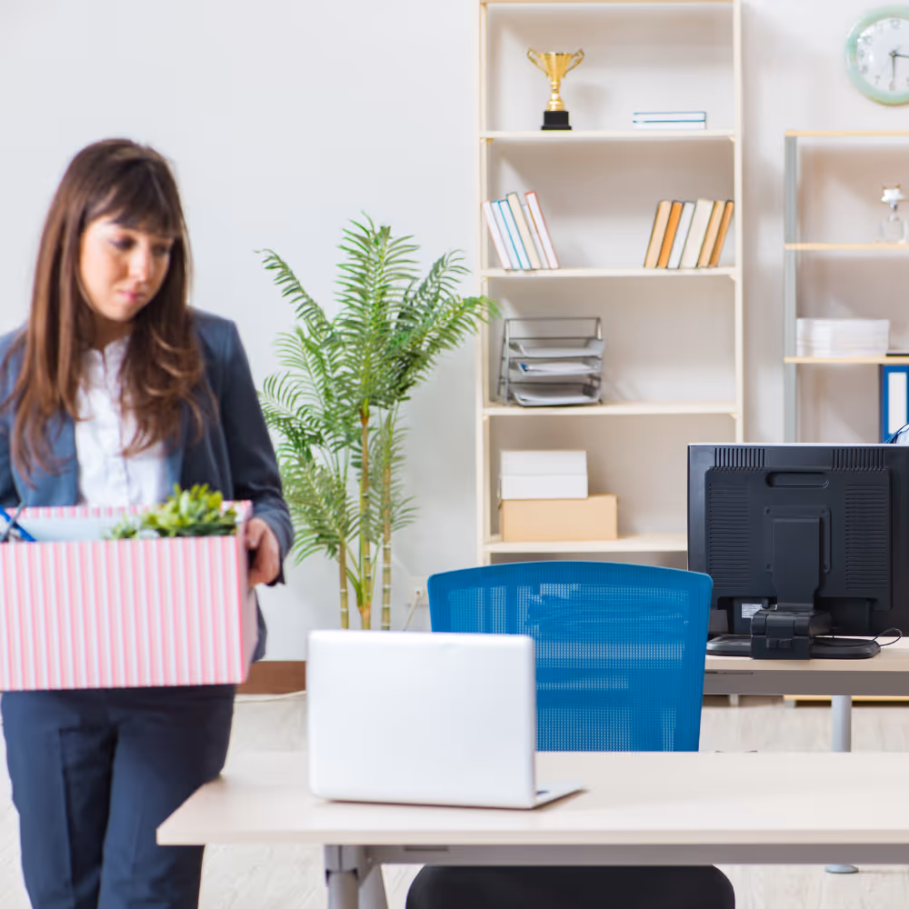 Woman carrying a striped box with office supplies, walking past a desk with a laptop in an office.