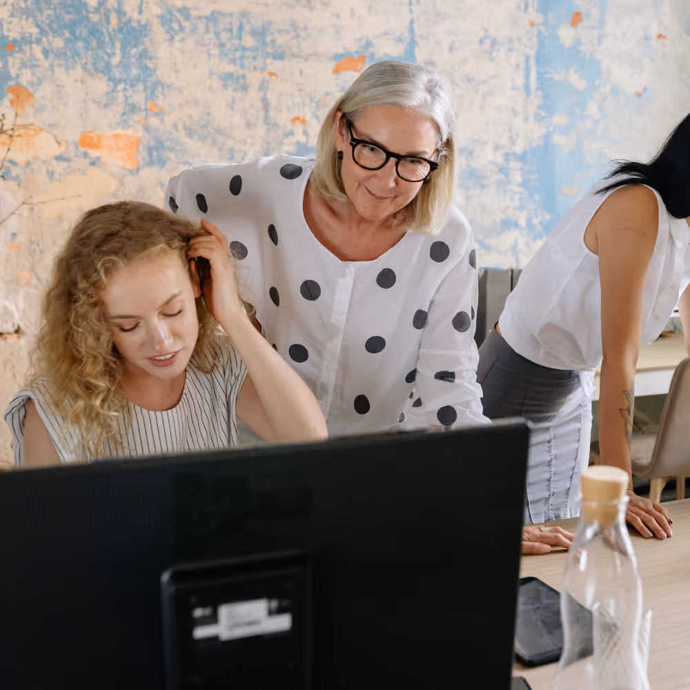 Two women looking at a computer screen with another person leaning on a table in the background.