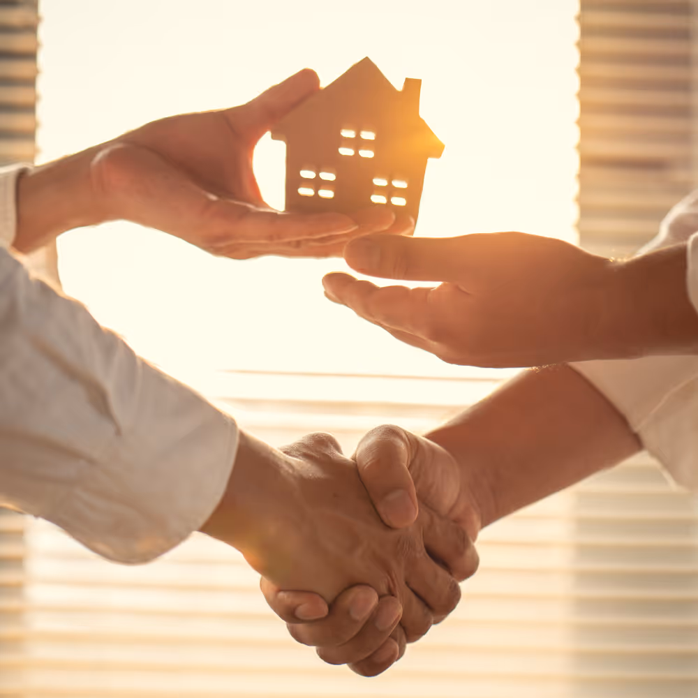 Two people shaking hands while holding a small wooden house shape between them.