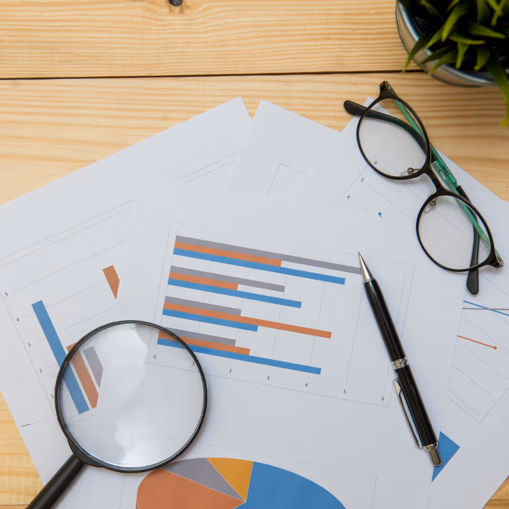 Magnifying glass, pen, and glasses on printed charts with bar graphs and pie charts on wood table.