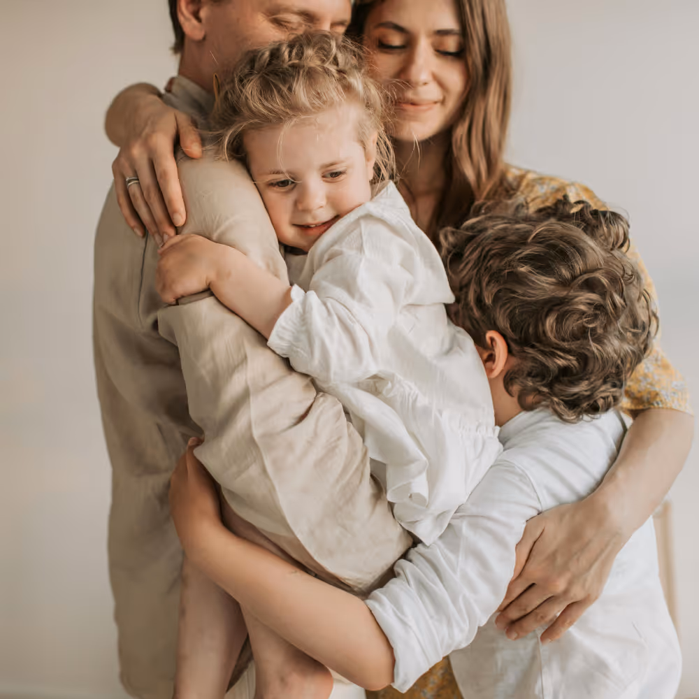 A family of four sharing a warm group hug with smiles and closed eyes in a soft, neutral setting.