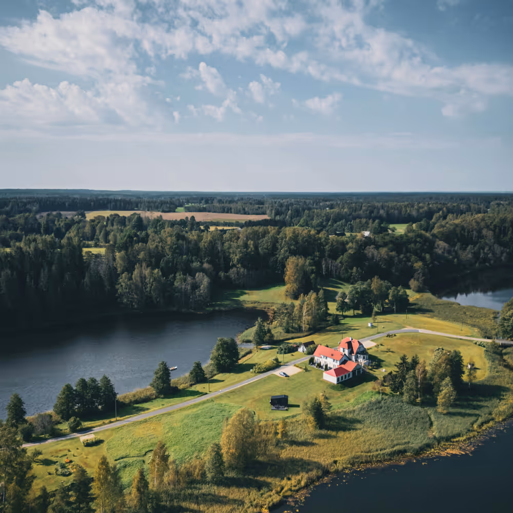 Aerial view of houses with red roofs surrounded by green fields, trees, and water bodies under a cloudy sky.
