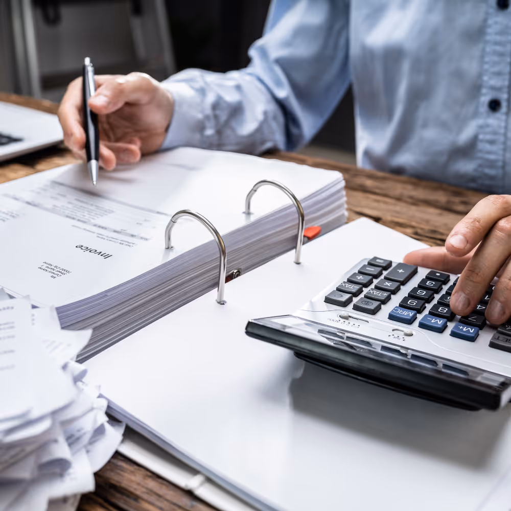 Person using a calculator and pen to review and organize invoices in a binder on a wooden table.