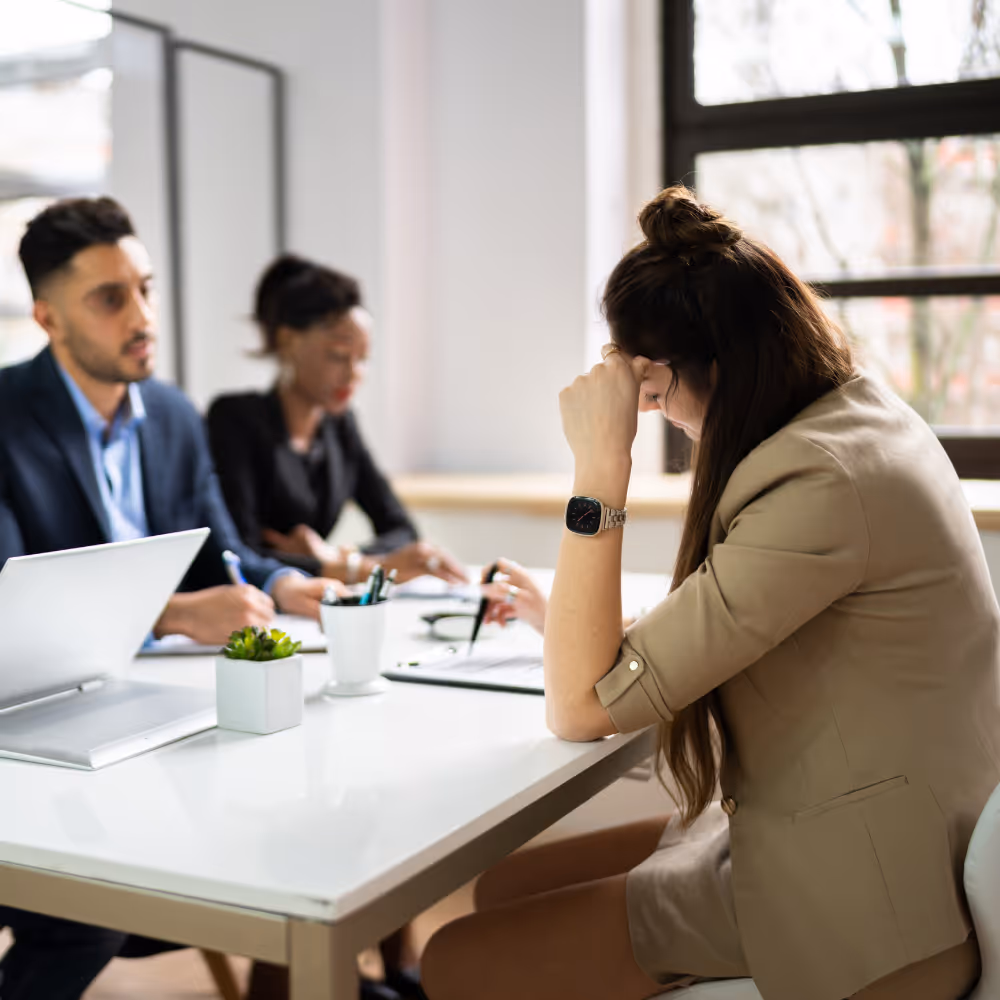 Stressed woman sitting at table holding head, with two blurred people in business attire in background.