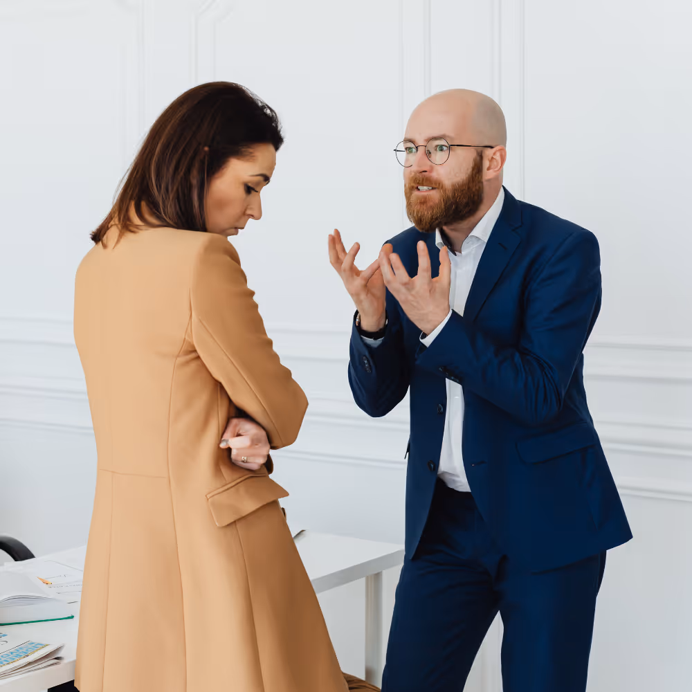 Man in blue suit gestures with hands talking to woman in beige coat who looks down, arms crossed.