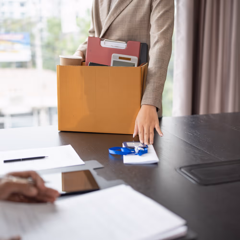 Person holding a box with office supplies, placing an envelope with ID badge on a table.