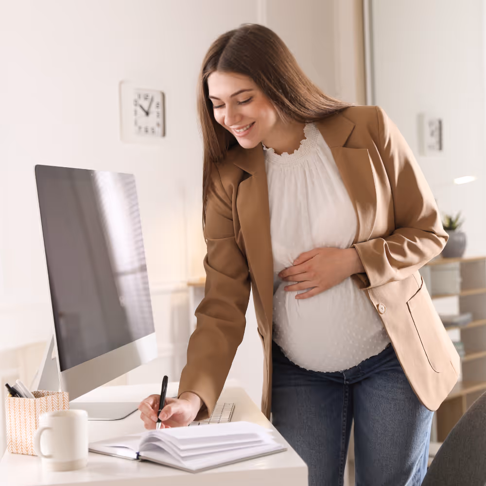 Pregnant woman in brown blazer writing in a notebook at her desk with a computer and mug.